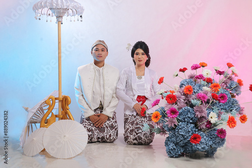 full length of a young Asian male and female couple sitting formally on the floor wearing modern Javanese traditional clothing, white velvet bridal kebaya on a white background. advertising content photo