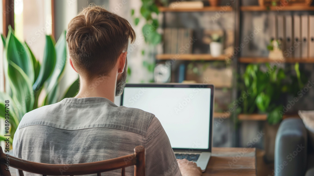 Over-the-shoulder shot of a young man focused on his laptop with a blank white screen, seated in a modern home office with a blend of contemporary and cozy decor. Over-the-shoulder shot of a young man focused on his laptop with a blank white screen, seated in a modern home office with a blend of contemporary and cozy decor.
