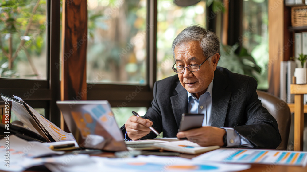 Obraz premium A senior middle-aged Asian businessman manager CEO, surrounded by documents and charts, uses his smartphone and laptop to streamline online business processes.