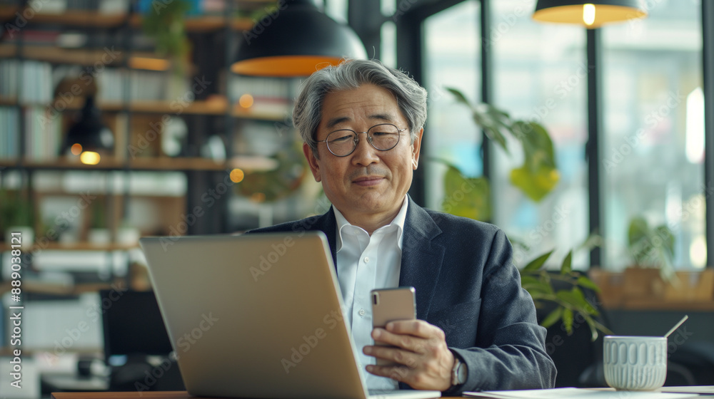 In a vibrant office setting, a senior middle-aged Asian businessman manager CEO uses a smartphone and laptop, symbolizing the dynamic and fast-paced nature of modern business manag In a vibrant office setting, a senior middle-aged Asian businessman manager CEO uses a smartphone and laptop, symbolizing the dynamic and fast-paced nature of modern business manag