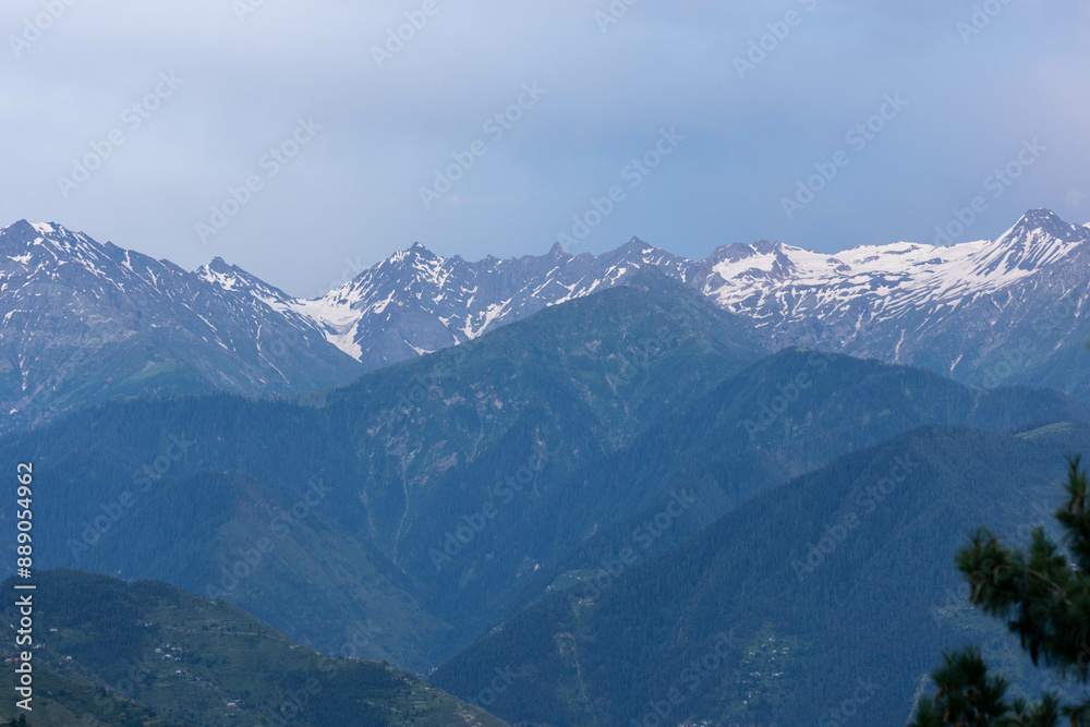 Snow-covered mountain range with cloudy skies and lush green valleys below Snow-covered mountain range with cloudy skies and lush green valleys below