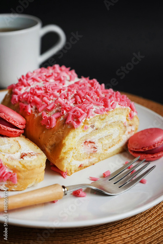 Sponge roll with cream, pink sprinkles and raspberry macaroons on a white plate next to a fork and cup on a dark background