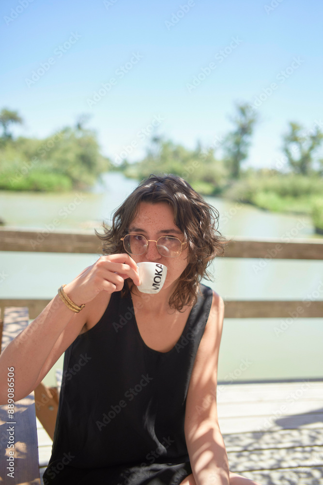 A person sits outside at a wooden table sipping from a coffee cup with a scenic water background and sunny weather. A person sits outside at a wooden table sipping from a coffee cup with a scenic water background and sunny weather.