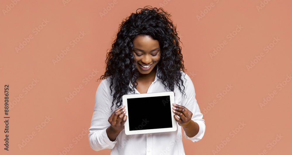A young black woman with curly black hair smiles brightly as she holds up a white tablet computer with a black screen in front of a plain peach background A young black woman with curly black hair smiles brightly as she holds up a white tablet computer with a black screen in front of a plain peach background