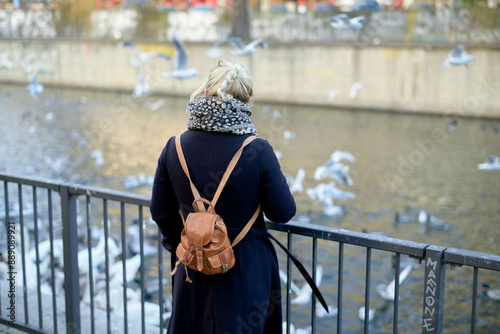 A woman stands by a railing overlooking a river filled with numerous birds, with seagulls in flight and water in the background. photo