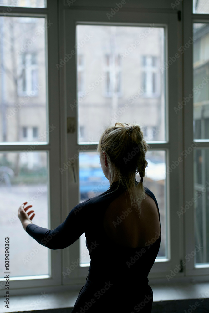 Back view of a woman with a ponytail standing by a window, gesturing with her hand, as she looks outside.