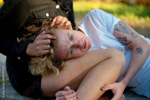 A person with curled blonde hair and tattoos resting their head on their knee outdoors, looking pensive.