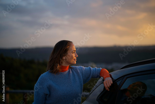 Woman leaning on a car door gazing into the distance during sunset.