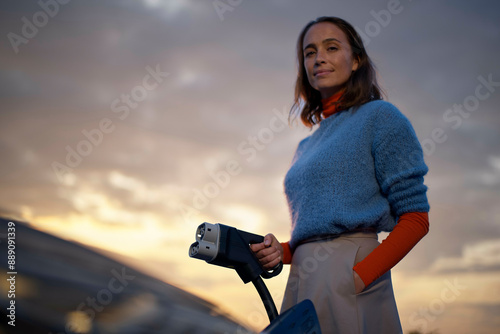 Confident woman holding an electric vehicle charging plug with a sunset sky in the background. photo