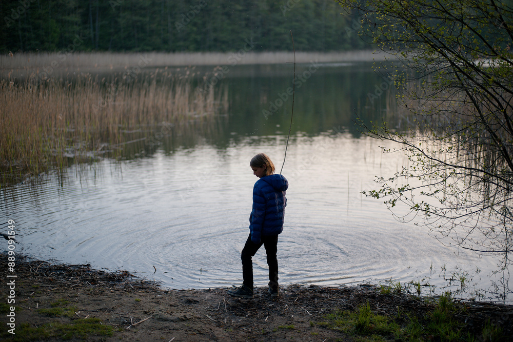 A child stands contemplatively at the edge of a serene lake surrounded by forest and reeds at dusk. A child stands contemplatively at the edge of a serene lake surrounded by forest and reeds at dusk.