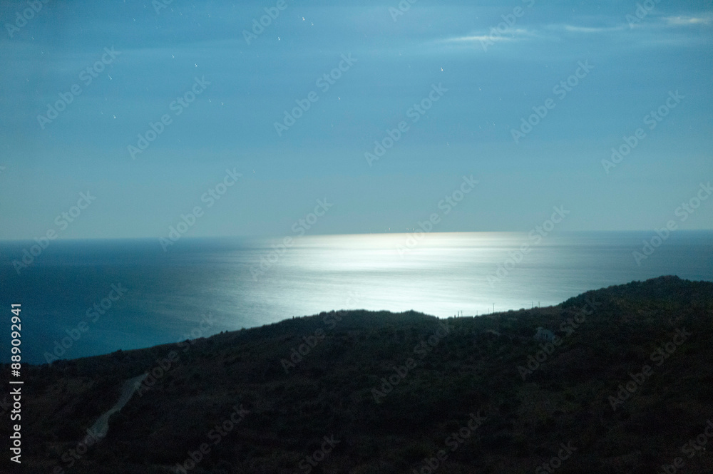 Moonlight reflects over a calm sea beyond a silhouetted coastline under a starry night sky. Moonlight reflects over a calm sea beyond a silhouetted coastline under a starry night sky.