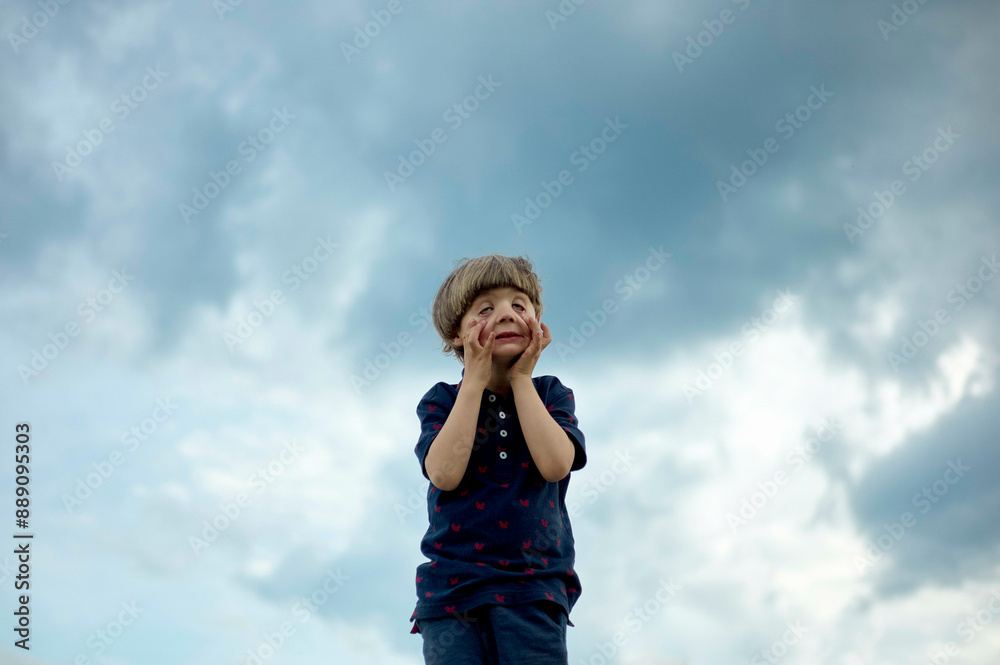 Young child standing against a cloudy sky, looking upwards with hands on cheeks in a gesture of surprise or awe. Young child standing against a cloudy sky, looking upwards with hands on cheeks in a gesture of surprise or awe.
