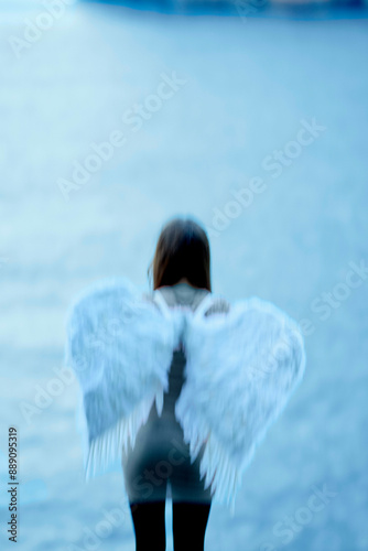 A blurred figure with angel wings stands facing away from the camera against a blue backdrop. photo