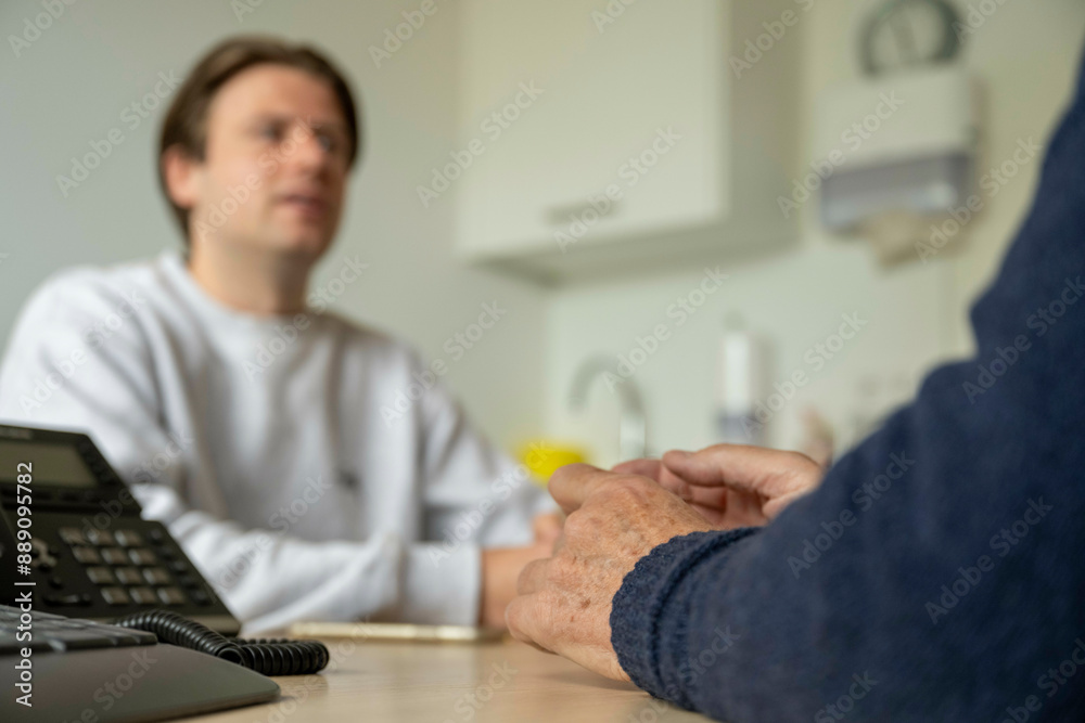 A doctor in a white coat sits across from a patient, hands clasped on a table with a landline phone in the foreground. A doctor in a white coat sits across from a patient, hands clasped on a table with a landline phone in the foreground.