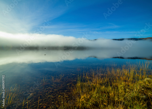 A serene lake reflects the mist and blue sky at sunrise, with golden grasses in the foreground. Loch Arkaig  photo