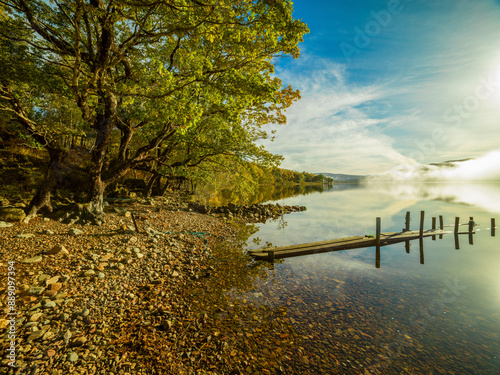 Serene lakeside view with a misty horizon, surrounded by trees with autumn foliage and a tranquil mirror-like water surface.