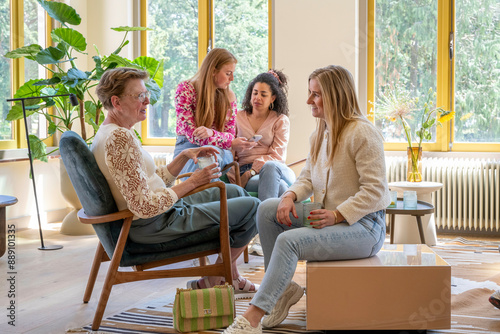 Four women of varying ages enjoying a casual conversation in a sunny living room, with large windows and lush greenery outside.
