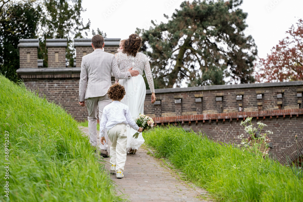 Bridal couple and young child walking hand in hand on a path in a park, dressed in wedding attire with the child carrying a bouquet. Bridal couple and young child walking hand in hand on a path in a park, dressed in wedding attire with the child carrying a bouquet.