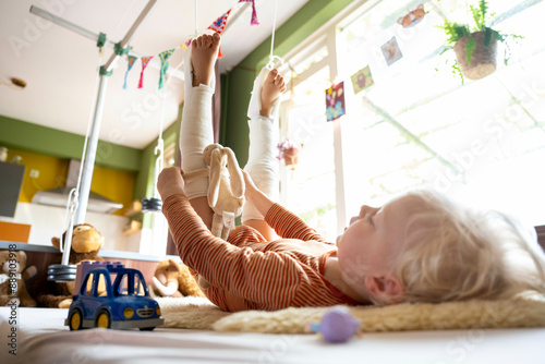 A child with leg casts plays with toys on a white blanket indoors, with plush toys in the background and a garden view through the window. photo