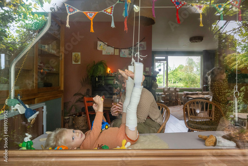 A toddler recovering from an injury with cast on his legs. plays with a hanging toy by a window with colorful decorations as a woman looks on from the cozy room inside. photo