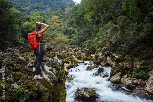 A hiker with a red backpack stands on a rocky outcrop overlooking a rushing river in a lush forest.