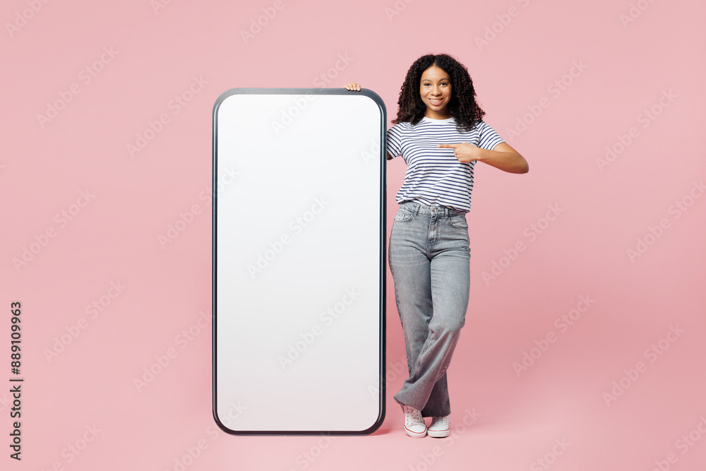 Full body little kid teen girl wear blue t-shirt casual clothes point finger on big huge blank screen mobile cell phone smartphone with area isolated on plain pastel pink background Childhood concept Full body little kid teen girl wear blue t-shirt casual clothes point finger on big huge blank screen mobile cell phone smartphone with area isolated on plain pastel pink background Childhood concept