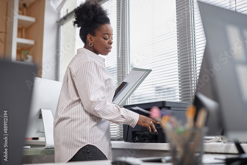 Medium shot of young female African American employee or student in casual white shirt using printer in office or class, copy space photo