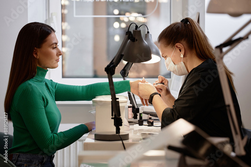 Side view shot of female nail tech wearing protective mask while doing manicure for young brunette woman during pampering beauty treatment in nail salon photo