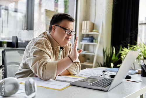 A young man with Down syndrome sits at a desk, waving at a laptop screen.