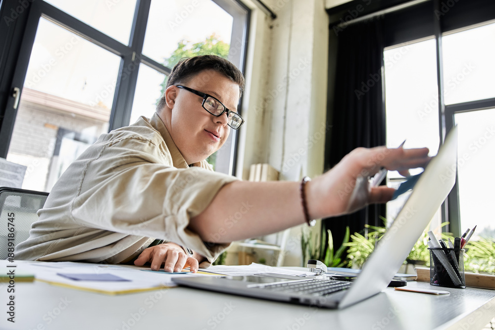 A young man with Down syndrome uses a laptop in a well lit office space. A young man with Down syndrome uses a laptop in a well lit office space.