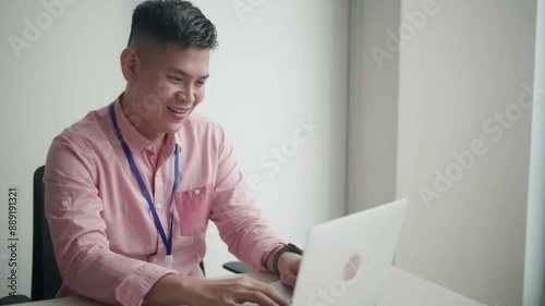 An Asian man in a pink shirt is happily working on his laptop at the office, smiling as he types, representing a positive work environment and job satisfaction in a modern workspace