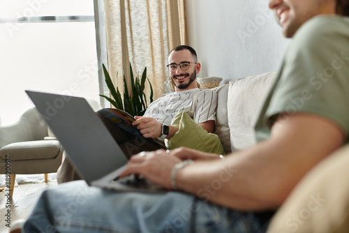 Couple relaxes on couch, one works on laptop while other reads magazine, sharing glance