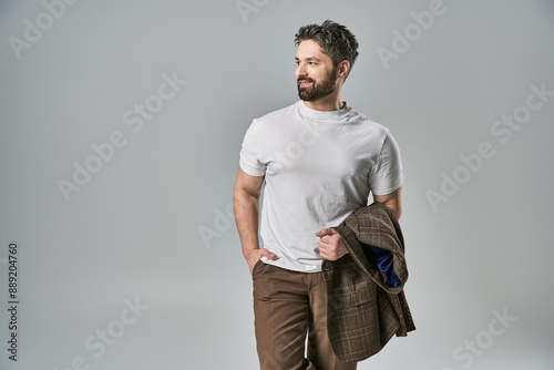 A stylish man with a beard poses confidently in elegant white t-shirt and brown pants against a grey studio background. photo