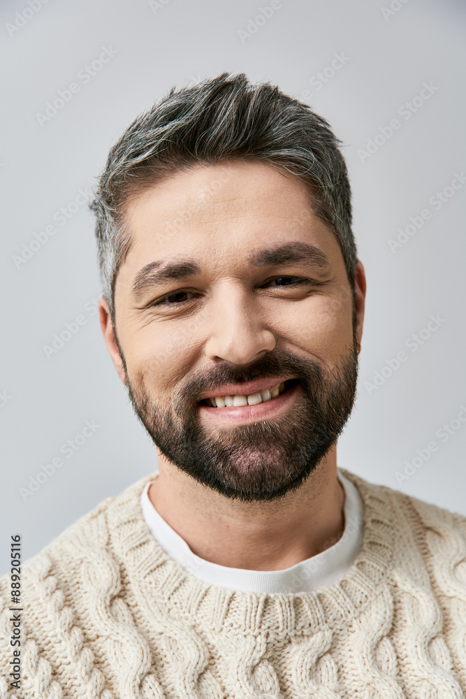 Fototapeta premium A serene, grey-haired man with a beard, wearing a white sweater, smiles warmly against a grey studio backdrop.