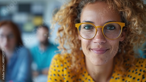 A smiling woman with curly hair wearing yellow glasses and a yellow polka-dotted shirt, prominently in focus while the background shows blurred figures in an indoor setting.