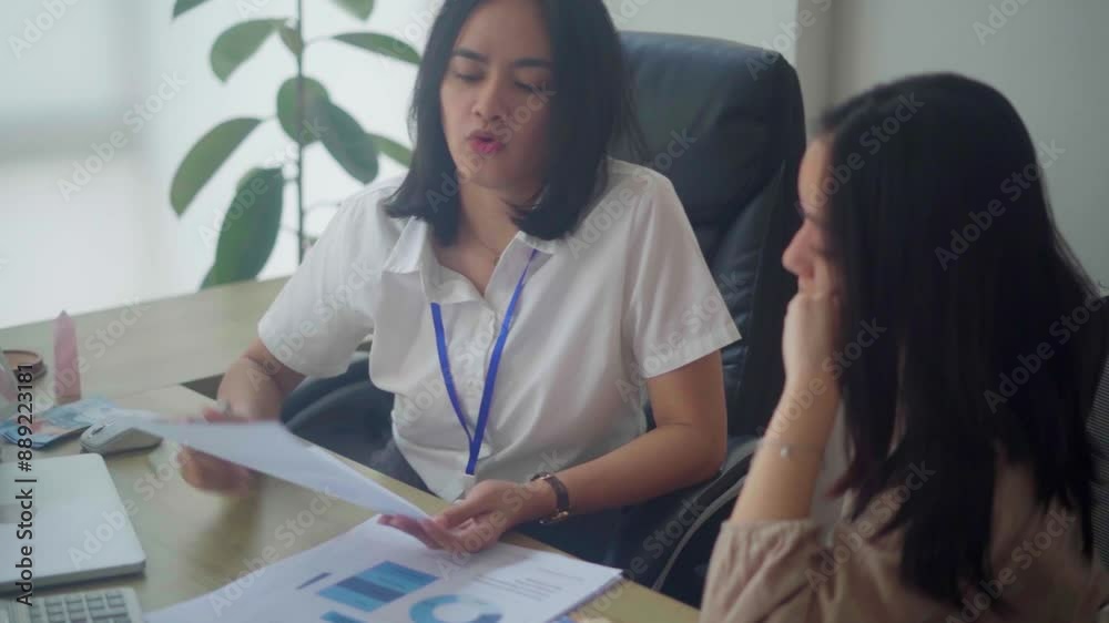 Two Asian women collaborating at a desk, discussing and reviewing documents in a modern office setting, focused on work-related tasks and decision-making. Two Asian women collaborating at a desk, discussing and reviewing documents in a modern office setting, focused on work-related tasks and decision-making.