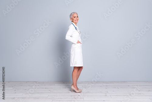 Full size side profile photo of therapist woman wear white uniform stethoscope on neck arms in pockets isolated on gray color background photo