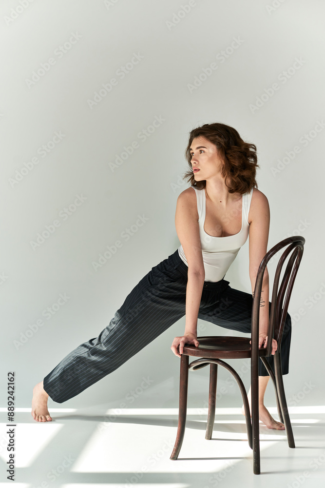 A pretty young woman in black pants and white tank top sits gracefully atop a wooden chair in a studio setting against a grey background. A pretty young woman in black pants and white tank top sits gracefully atop a wooden chair in a studio setting against a grey background.