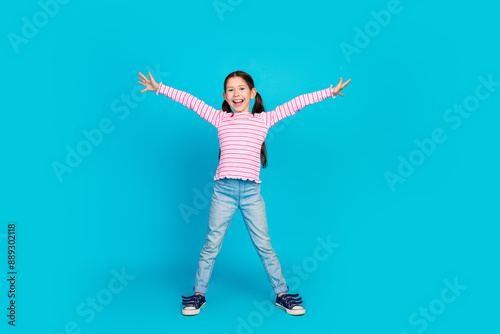 Full size photo of carefree funky small girl with ponytails dressed striped shirt raising hands up isolated on blue color background