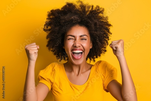 Black woman with fist celebrating promotion or lottery bonus on yellow background. Happy model, deal, or lottery winner in studio