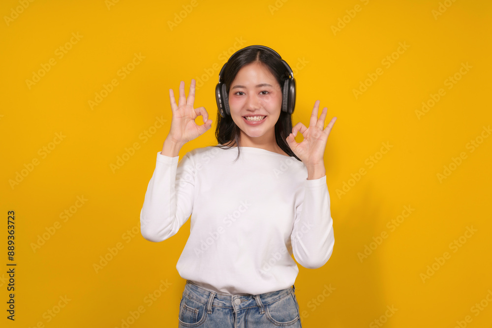 Young woman is enjoying music on her headphones while giving the ok sign with a smile, isolated on a vibrant yellow background Young woman is enjoying music on her headphones while giving the ok sign with a smile, isolated on a vibrant yellow background