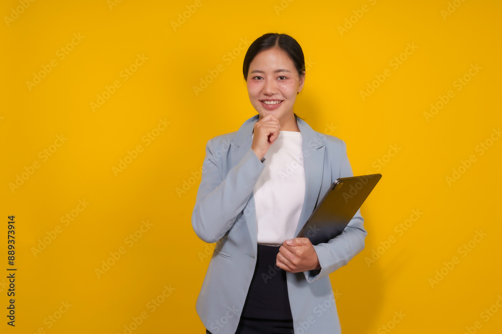 Young asian businesswoman is posing with a tablet in her hand, smiling and thoughtfully touching her chin on a vibrant yellow background Young asian businesswoman is posing with a tablet in her hand, smiling and thoughtfully touching her chin on a vibrant yellow background