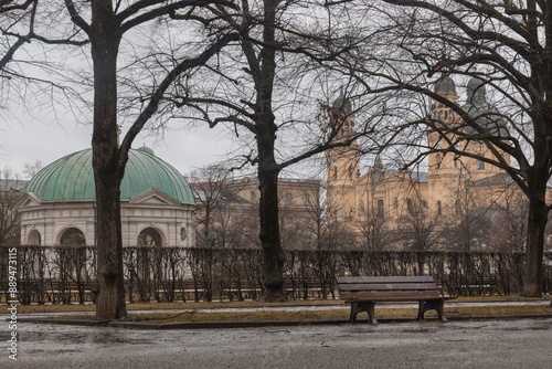 Munich, Germany - Dec 22, 2023 - Bench made wood and iron stand in the garden of the Munich Residenz with The Diana Temple (Dianatempel) and St.Cajetan and Adelaide in the background. Space for text,  photo