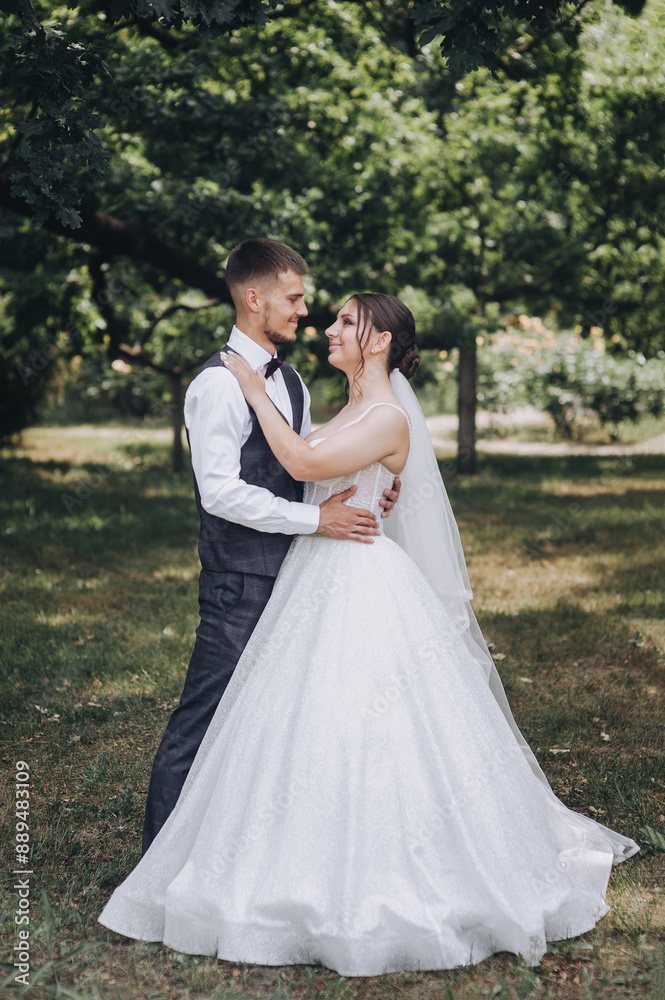 Beautiful young happy smiling groom in a gray suit and a bride in a white dress with a bouquet of flowers stand hugging outdoors in the park. Wedding photography, portrait of stylish newlyweds. Beautiful young happy smiling groom in a gray suit and a bride in a white dress with a bouquet of flowers stand hugging outdoors in the park. Wedding photography, portrait of stylish newlyweds.