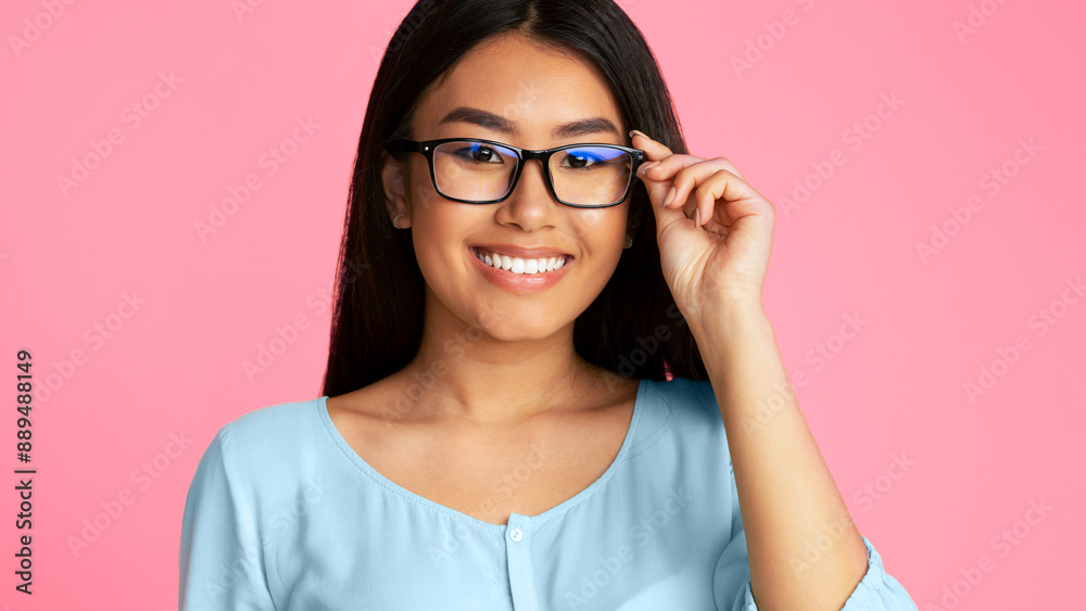 Asian young woman with long black hair smiling while wearing eyeglasses against a pink background. She is wearing a light blue blouse, holding the right side of her glasses with her right hand. Asian young woman with long black hair smiling while wearing eyeglasses against a pink background. She is wearing a light blue blouse, holding the right side of her glasses with her right hand.