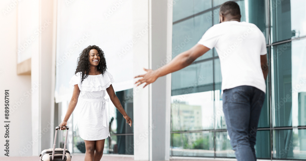 Black woman with curly hair is pulling a rolling suitcase and smiling at a man walking towards her with his arms outstretched. They are both standing in an airport terminal. Black woman with curly hair is pulling a rolling suitcase and smiling at a man walking towards her with his arms outstretched. They are both standing in an airport terminal.