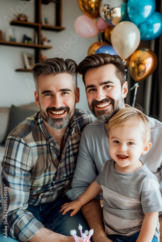 vertical portrait of happy gay couple dads with adopted toddler baby celebrating birthday at camera at home