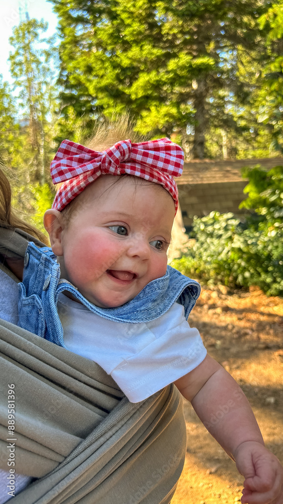 Caucasian Baby Girl Wearing Denim Overalls and Red Bow Smiling in Baby Carrier Outside Caucasian Baby Girl Wearing Denim Overalls and Red Bow Smiling in Baby Carrier Outside