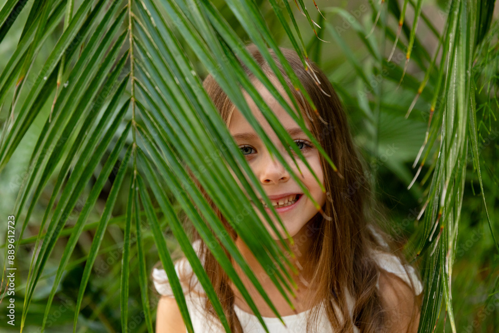 Fototapeta premium The face of a happy little girl surrounded by tropical leaves. Closeup portrait of a beautiful child with perfect skin and dark hair. Natural cosmetics, health, cleanliness, skin care, beauty concept