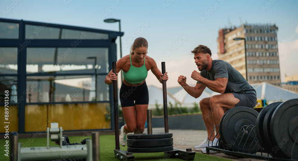 Sporty young woman pushes a weighted sled on artificial turf while a man cheers her on at an outdoor gym. Sporty young woman pushes a weighted sled on artificial turf while a man cheers her on at an outdoor gym.